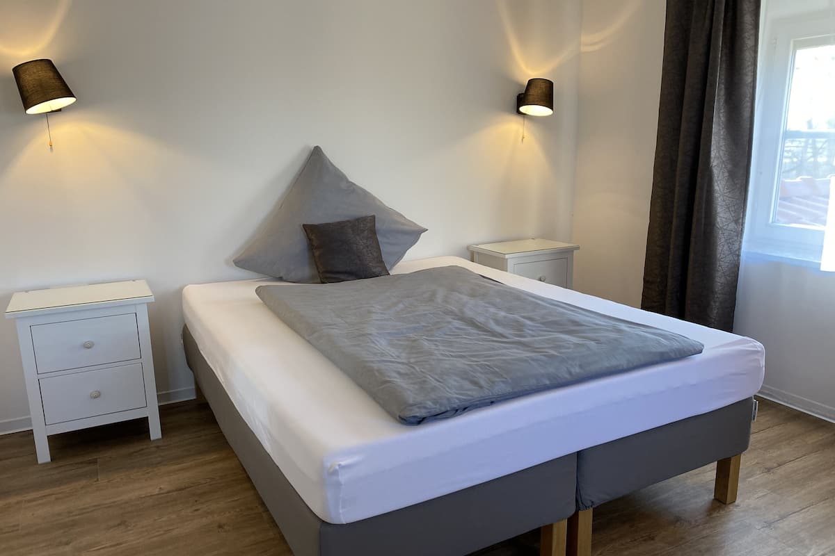 Quarto com cama, duas mesas de cabeceira, um criado-mudo, janela com cortinas e iluminação na parede.
