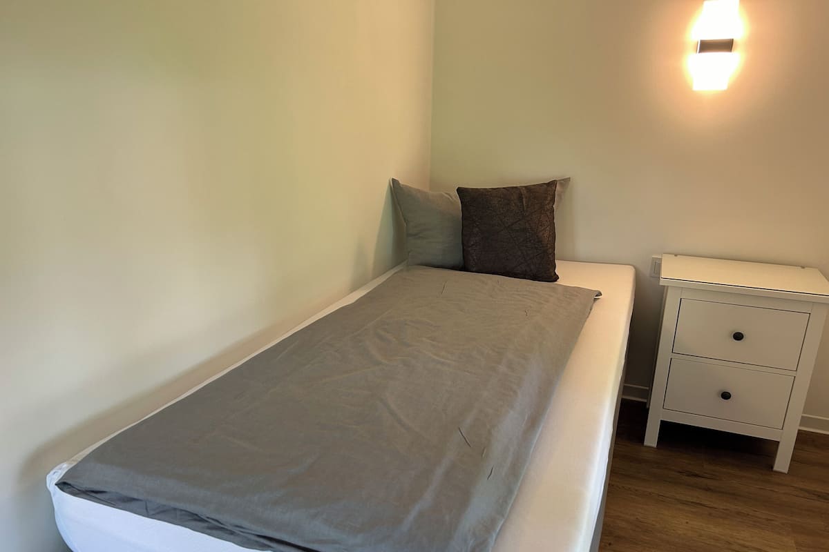 Uma cama de solteiro com um cobertor cinzento, um travesseiro branco e um travesseiro castanho sobre um colchão branco. Há uma mesinha de cabeceira branca com duas gavetas à direita da cama.