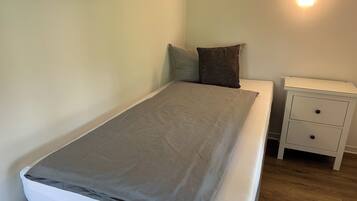 Ein Einzelbett mit grauer Decke, einem weißen Kopfkissen und einem braunen Kissen auf einer weißen Matratze. Rechts daneben ein Nachttisch in Weiß mit zwei Schubladen.