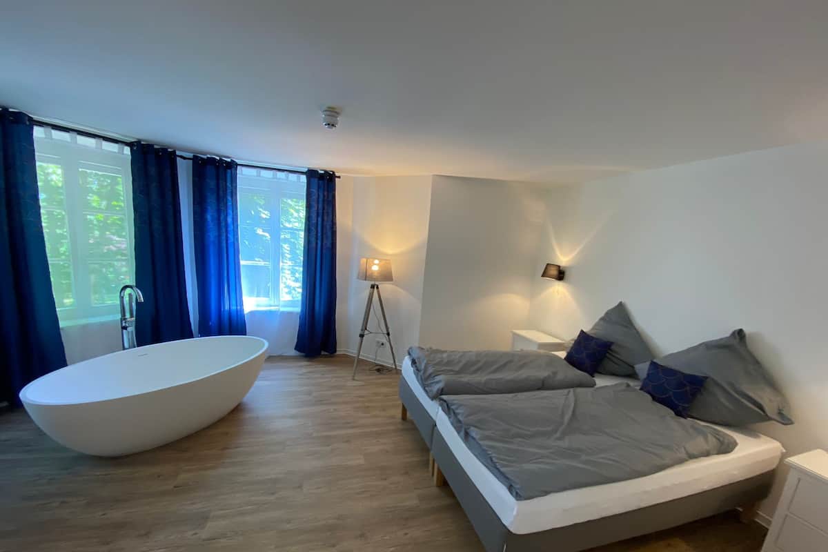 Quarto moderno com uma banheira grande, uma cama com roupa de cama cinza e um abajur.