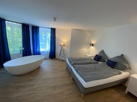 Quarto moderno com uma banheira grande, uma cama com roupa de cama cinza e um abajur.