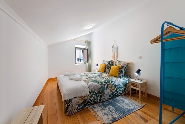 A bedroom with a bed, a small bedside table, a hanging light, a window, and a blue curtain.