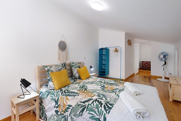 A bedroom with a bed, a bedside table, a mirror, a blue shelving unit, a fan, and a wooden chest of drawers.