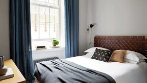 Un dormitorio ordenado con una cama grande, una mesita de noche con un jarrón y libros, una ventana con cortinas y una lámpara de pared.