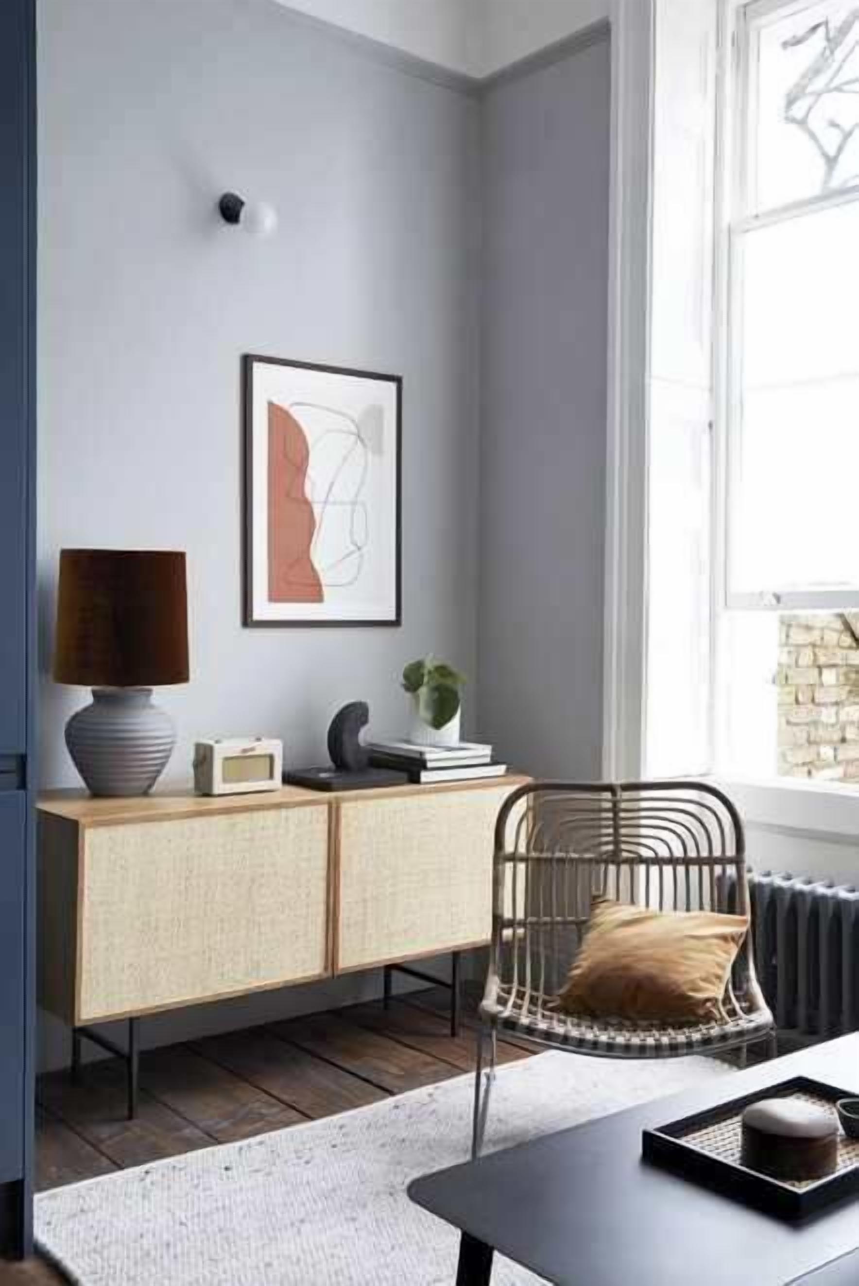 A modern living room with a grey wall, a framed abstract artwork, a wooden cabinet, a wicker chair, and a table with a tray.