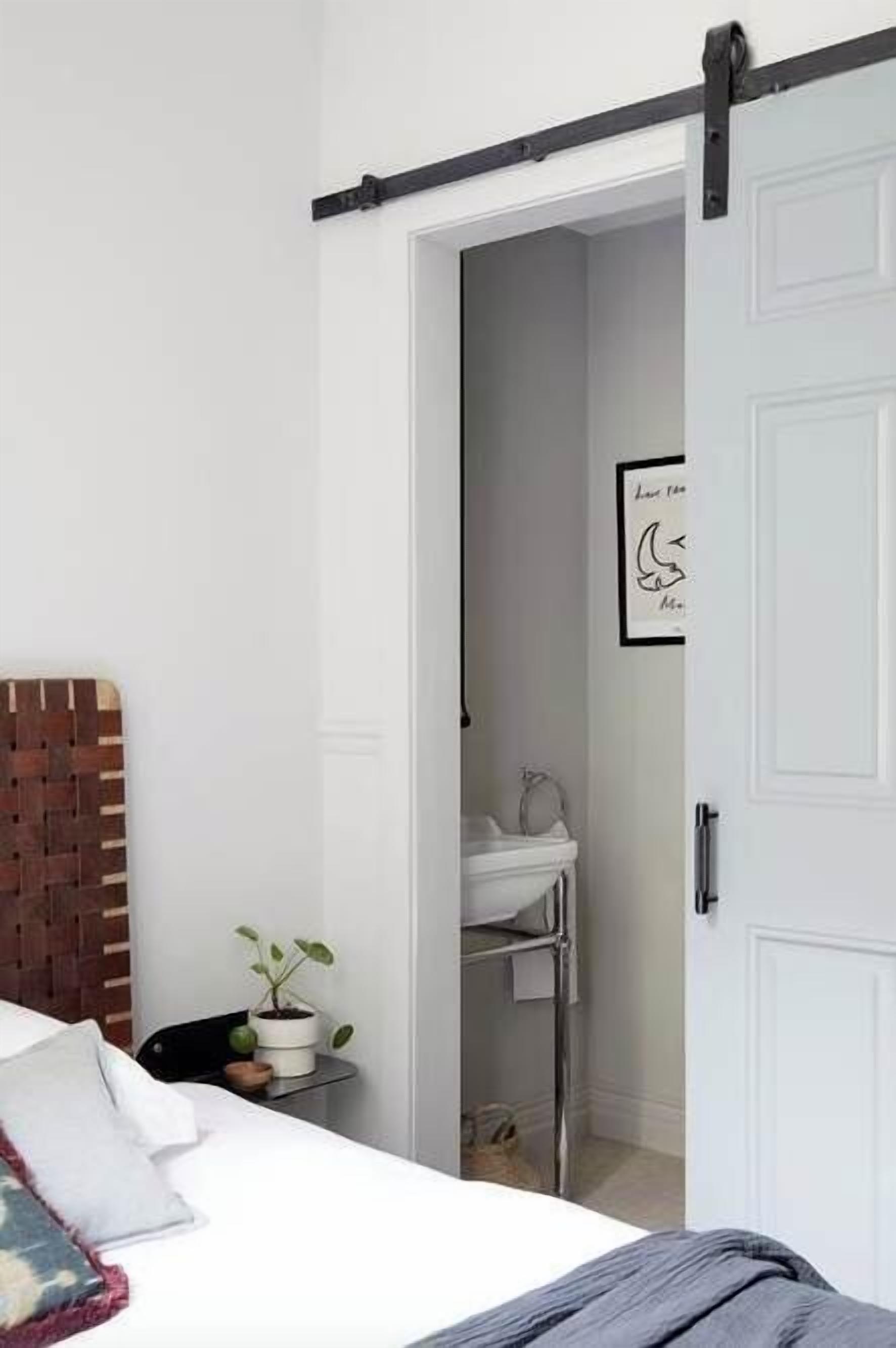 Ein Schlafzimmer mit Bett, Nachttisch mit Pflanze, weißer Tür und einem gerahmten Bild an der Wand.