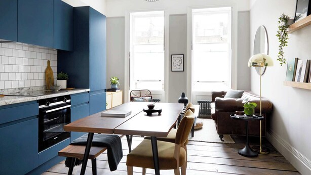 Una cocina moderna con armarios azules, una mesa de comedor de madera y un sofá en un espacio luminoso con amplias ventanas.