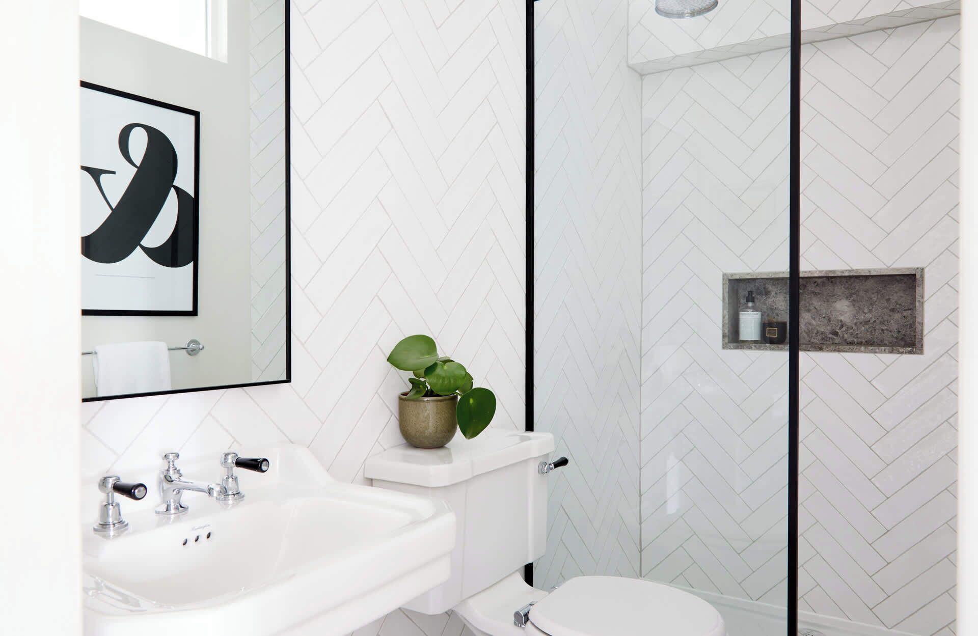 A modern bathroom with a white herringbone tile backsplash, a white sink, a toilet, and a glass-enclosed shower. There is a framed artwork above the sink and a potted plant on the sink's edge.
