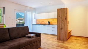 A modern living room with a sofa, wooden floor, and a kitchen with white cabinets and a wooden accent wall.