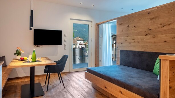 Una habitación moderna con una mesa de madera, un sofá, un televisor montado en la pared y un ventanal corredero que da a un balcón.