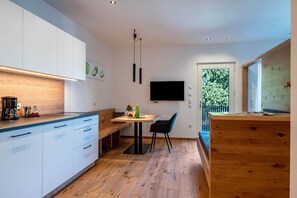 A modern kitchen with white cabinets, a wooden countertop, a dining table with chairs, a TV mounted on the wall, and a window with a view of greenery.