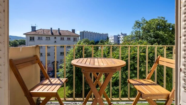 Un balcon avec deux chaises en bois et une petite table ronde, d’où l’on peut admirer un paysage urbain avec ses bâtiments et sa verdure.