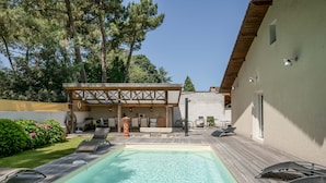 A pool area with lounge chairs, a covered seating area, and a building with a light-colored exterior. - Vacation Villa "Villa Teny La Teste Arcachon" with Pool, Garden, and Terrace (La Teste-de-Buch)