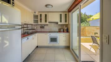 A compact kitchen with a refrigerator, oven, and sink, a sliding glass door to a balcony, and a chair outside.