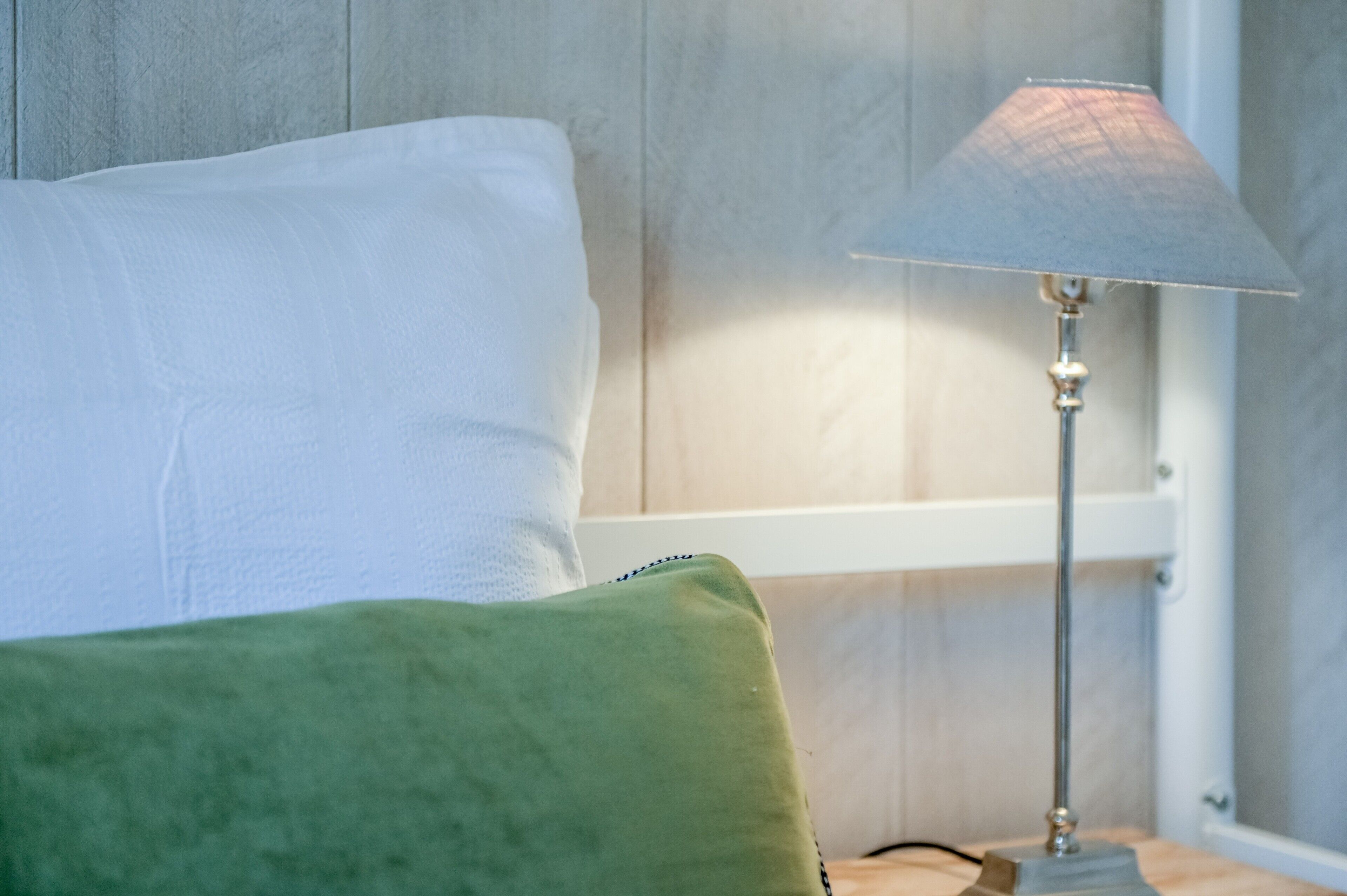 A bed with a white pillow and a green pillow, a lamp on a bedside table, and a wooden wall.