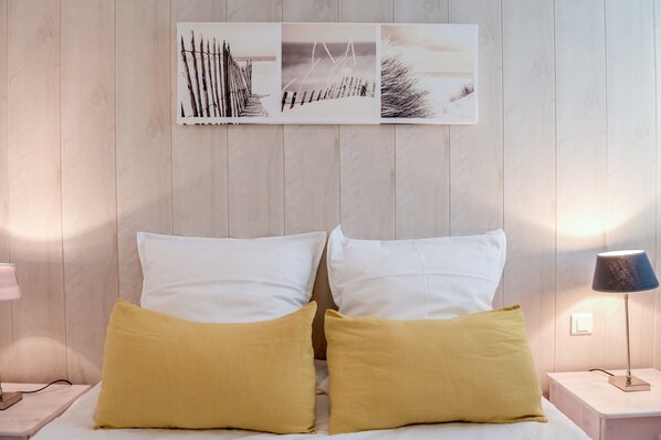 A bed with white and mustard pillows, a bedside lamp, and a wall-mounted picture of a fence and beach scene.