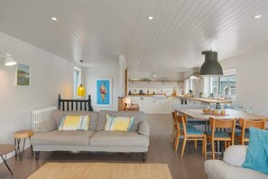A modern living room with a sofa, a dining table with chairs, a kitchen area, and a large window. - Stylish Home with Parking, Minutes from Beach (Bude)