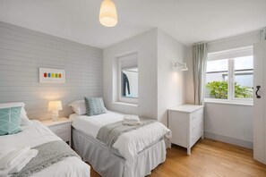 A bedroom with two beds, a bedside table with a lamp, a dresser, and a window with a view of greenery. - Stylish Home with Parking, Minutes from Beach (Bude)