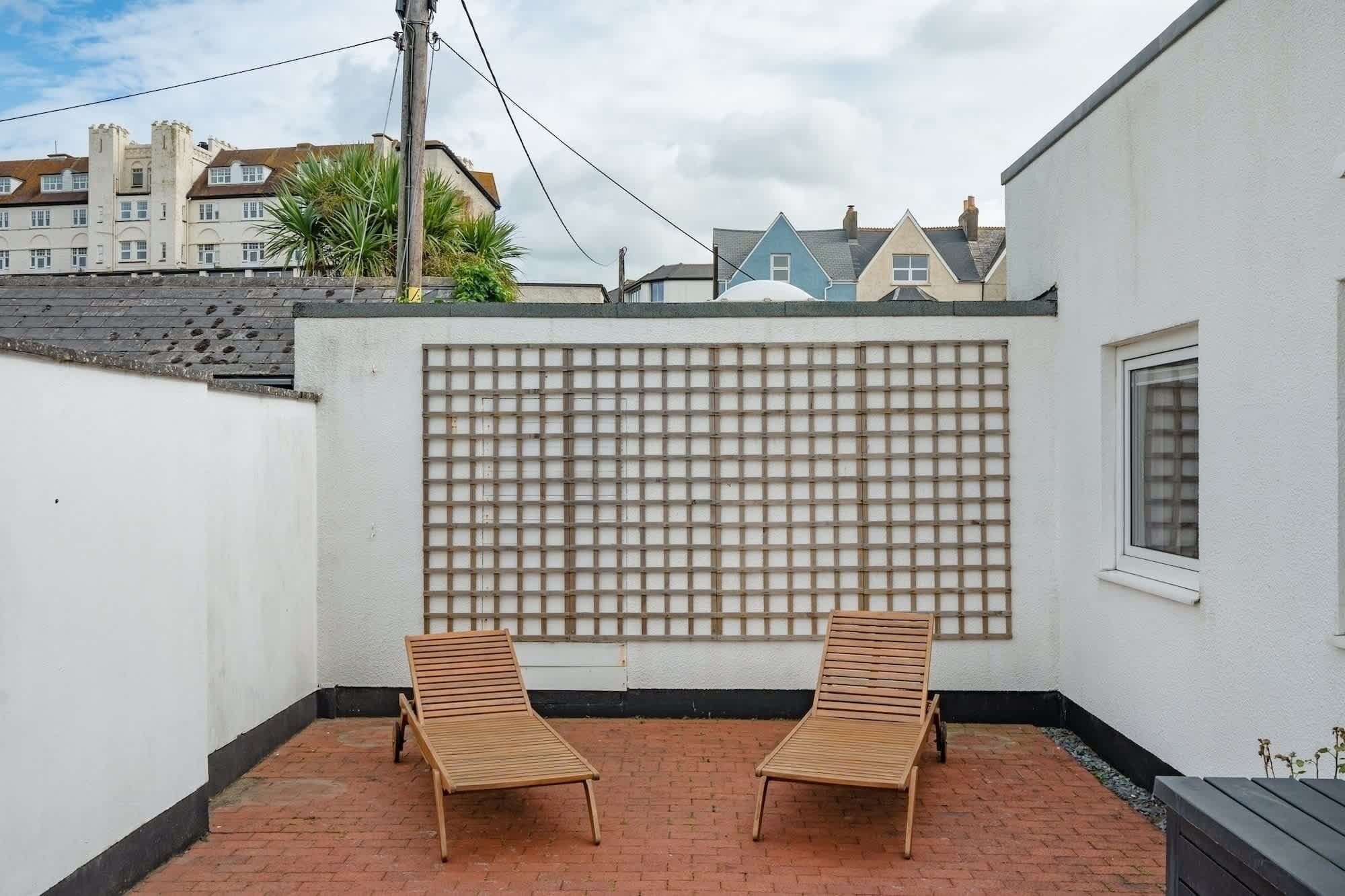 A courtyard with two wooden lounge chairs, a white wall with a grid pattern, and a view of neighboring houses.