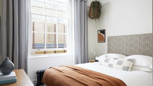 Ein Schlafzimmer mit großem Fenster, einem Bett mit gemusterten Kissen, einer hängenden Pflanze und einer Heizung.
