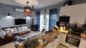 A modern kitchen with a built-in oven, a dining area with a wooden table, and a living room with a sofa and TV.
