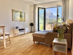 Une chambre moderne avec un plancher en bois, un lit recouvert d’une couverture beige, une table de chevet ornée d’un vase de fleurs, une commode blanche, un ventilateur sur pied et une grande fenêtre donnant sur les montagnes.