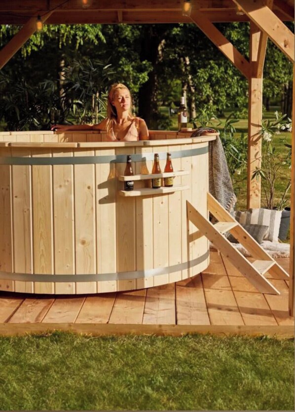 A wooden hot tub with a wooden deck, surrounded by greenery.