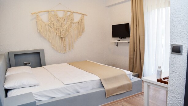 A bedroom with a bed, a TV, a macrame wall hanging, and a window with curtains.