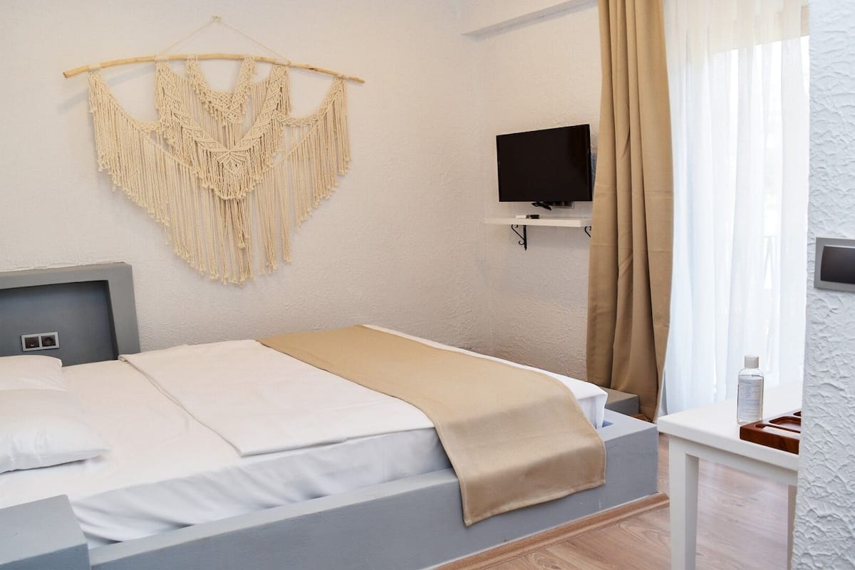Um quarto com cama, televisão, um painel de macramé e uma janela com cortinas.