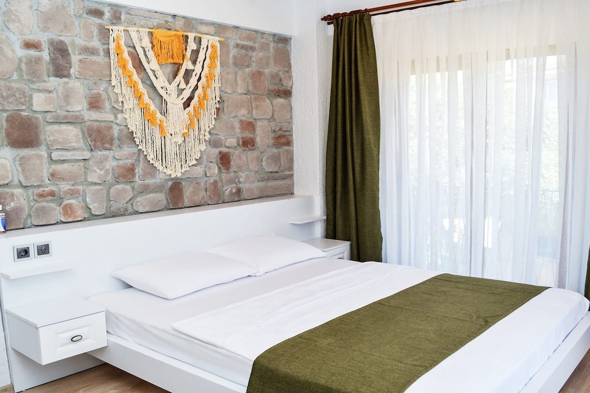 Quarto com parede de pedra, cama com roupa de cama branca, mesa de cabeceira com gaveta e janela com cortinas verdes.