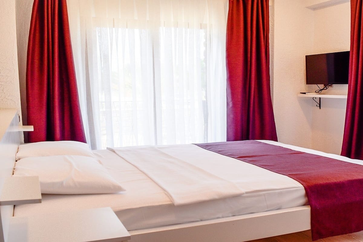 Quarto de hotel com cama, roupa de cama branca, manta vermelha, televisão na parede e cortinas vermelhas.