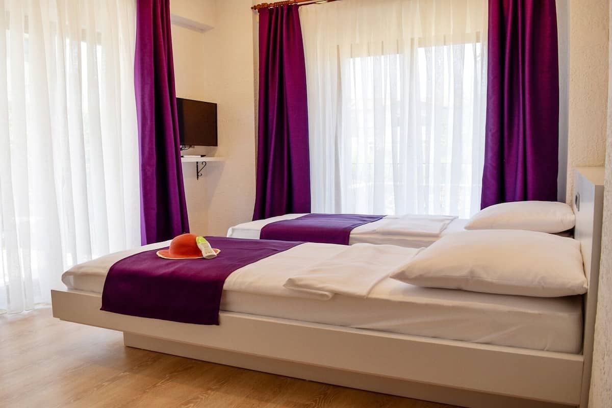 Quarto de hotel com cama, duas almofadas, cobertor, televisão e janela com cortinas.