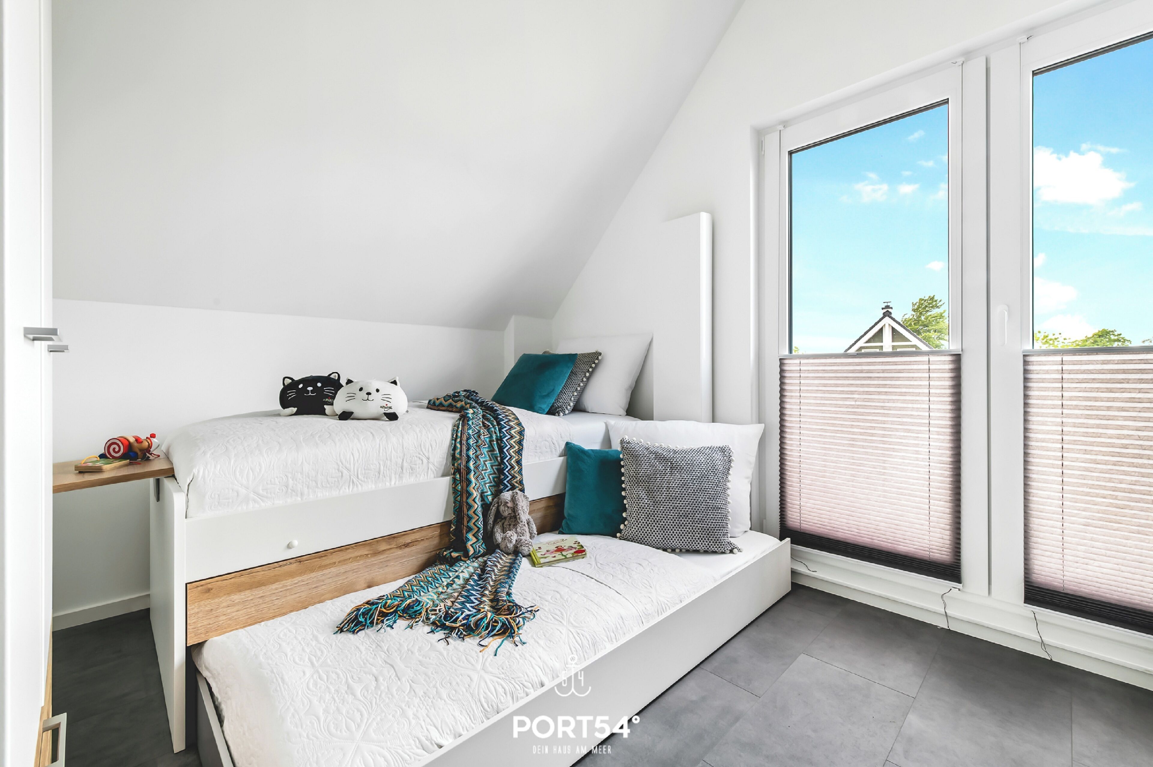 Ein modernes, gut beleuchtetes Zimmer mit einem Schlafplatz in der Mansarde, einem Schreibtisch und einem großen Fenster mit Blick auf Himmel und Gebäude.