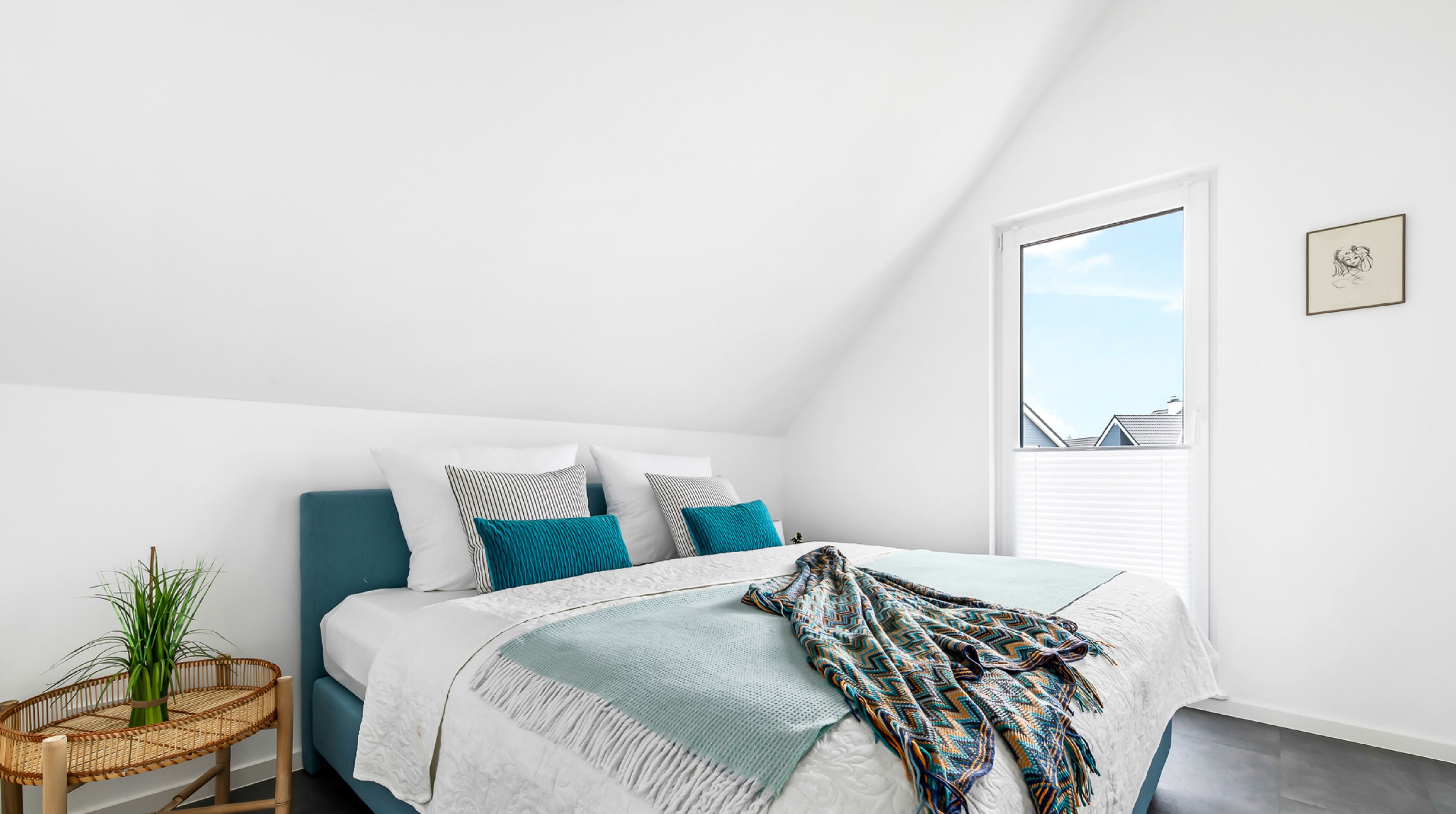 Ein Schlafzimmer mit Bett, Fenster, einem gerahmten Bild und einem kleinen Tisch mit Pflanze.