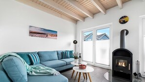A modern living room with a blue sofa, a fireplace, a painting of a beach, and a window with blinds. - Bajo Lykke in the North Sea resort - close to the beach and the North Sea, fireplace, infrared sauna, Terra (Büsum)