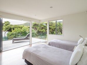 Two beds with white bedding, a sliding door to a deck with a lounge chair, and a view of lush greenery.