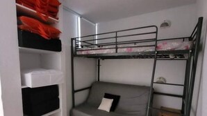Quarto com beliche, piso de madeira, uma escrivaninha pequena e prateleira com bolsas vermelhas.