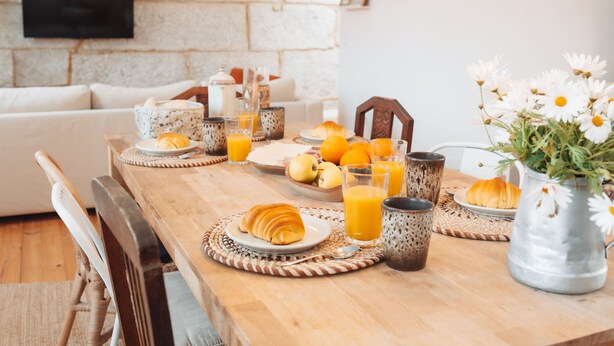 A breakfast table set with croissants, fruit, and orange juice, a vase of flowers, and a television mounted on a stone wall. - Biscoito Guest House (Viana do Castelo)