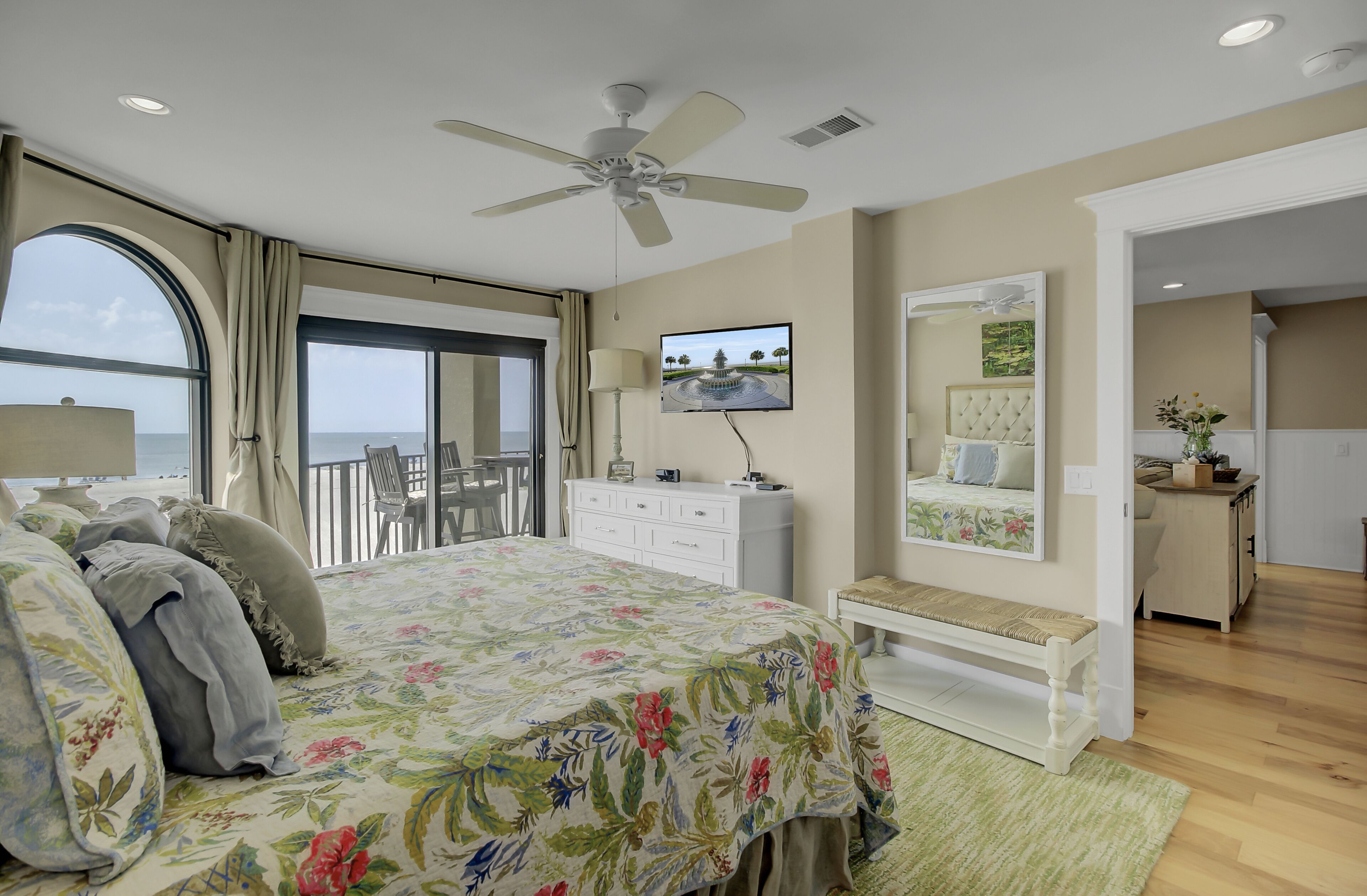 Une chambre à coucher comprenant un lit, une télévision, un ventilateur de plafond, une grande fenêtre avec des rideaux, une commode, un banc et une vue sur la mer.