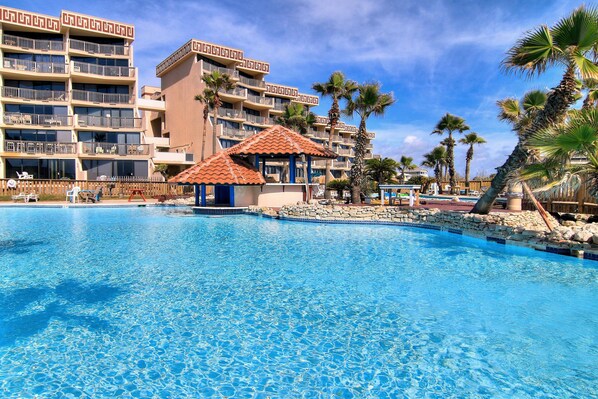 A swimming pool with a gazebo, palm trees, and a multi-story hotel building.