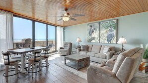 A spacious living room with a large window overlooking the ocean, a ceiling fan, and a comfortable seating area with a sofa and chairs. - Seaside Haven | Beachfront | Panoramic Ocean Views (Port Aransas)