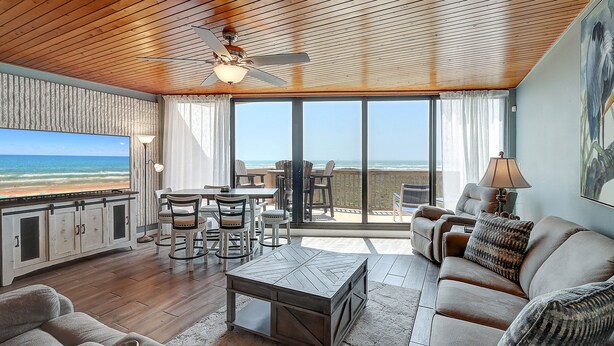 A spacious living area with a wooden ceiling, a large TV, a dining table, and a sofa. - Seaside Haven | Beachfront | Panoramic Ocean Views (Port Aransas)