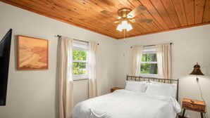 A bedroom with a wooden ceiling, a bed with white bedding, two windows with curtains, a nightstand with a lamp, and a framed picture on the wall.