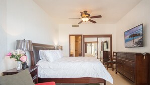 Une chambre à coucher comprenant un lit, une table de chevet, une commode, un ventilateur de plafond, un tableau et une vue sur la plage par une porte ouverte.