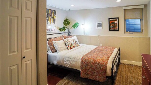 A bedroom with a bed, a nightstand, a dresser, a window with a view, a plant, and artwork on the wall.