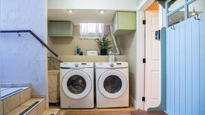 A compact laundry room with two front-loading washing machines, a small sink, and a window with a curtain.