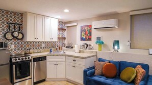 Cocina compacta con armarios blancos, estufa y lavavajillas. Hay un sofá azul con cojines coloridos y un cuadro en la pared.