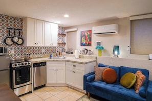 A compact kitchen with white cabinets, a stove, and a dishwasher. There's a blue sofa with colorful cushions and a painting on the wall.