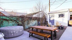 Un patio con mesa y banco de madera, rodeado por una valla verde y un edificio blanco.