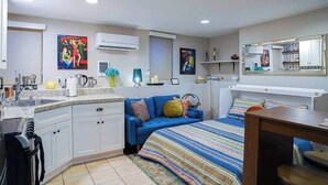 Una habitación pequeña con kitchenette, una cama con manta a rayas, un sofá azul y una mesita.
